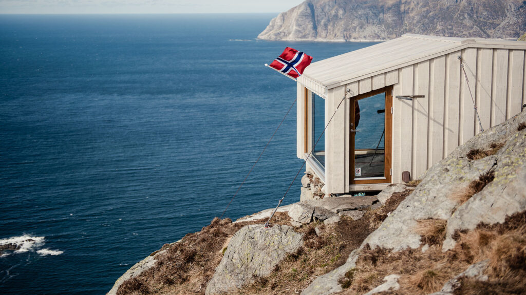 Eit lyst og moderne treskur er plassert på ein fjellklippe. Den har store glas ut mot havet og horisonten. Det er eit blafrande noregsflagg på sjøsida av bua. Foto.
