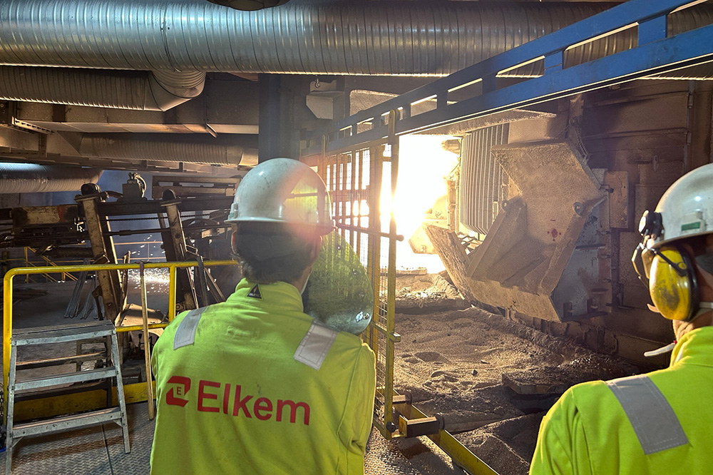 To personar i neongule arbeidsklede og tryggleiksutstyr ser mot eit kraftig lys i bakgrunnen. Uniformen ber logoen til Elkem. Foto.