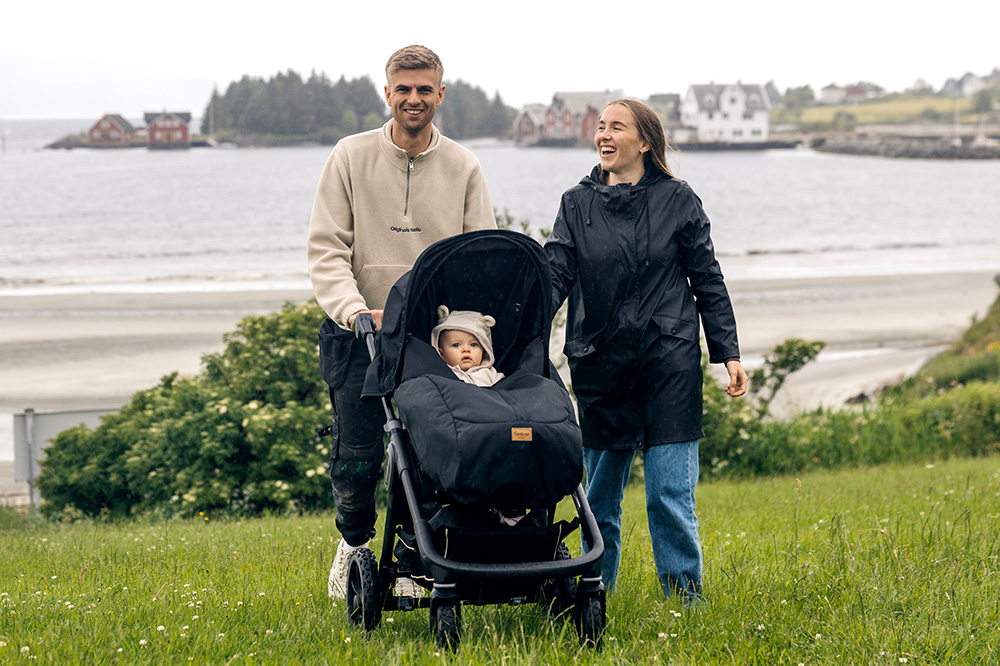 Mann og kvinne ler og triller ei barnevogn over graset. Babyen er våken og kikker ut mot kamera. Foto.