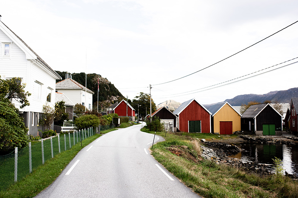 Ein tynn snirklete veg svingar rundt fargerike naust og kvite hus. Leidningane frå lyktestolpene følgjer vegen. Foto.