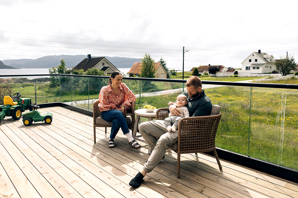 Eit par sit i kvar sin stol på ein veranda, og smiler ned på babyen deira som sit i far sitt fang. I bakgrunnen er det fleire grøne leiketraktorar. Foto.