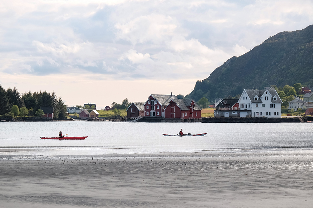 To personar i kvar sin kayak padlar forbi eit stort tradisjonelt naust. Foto.