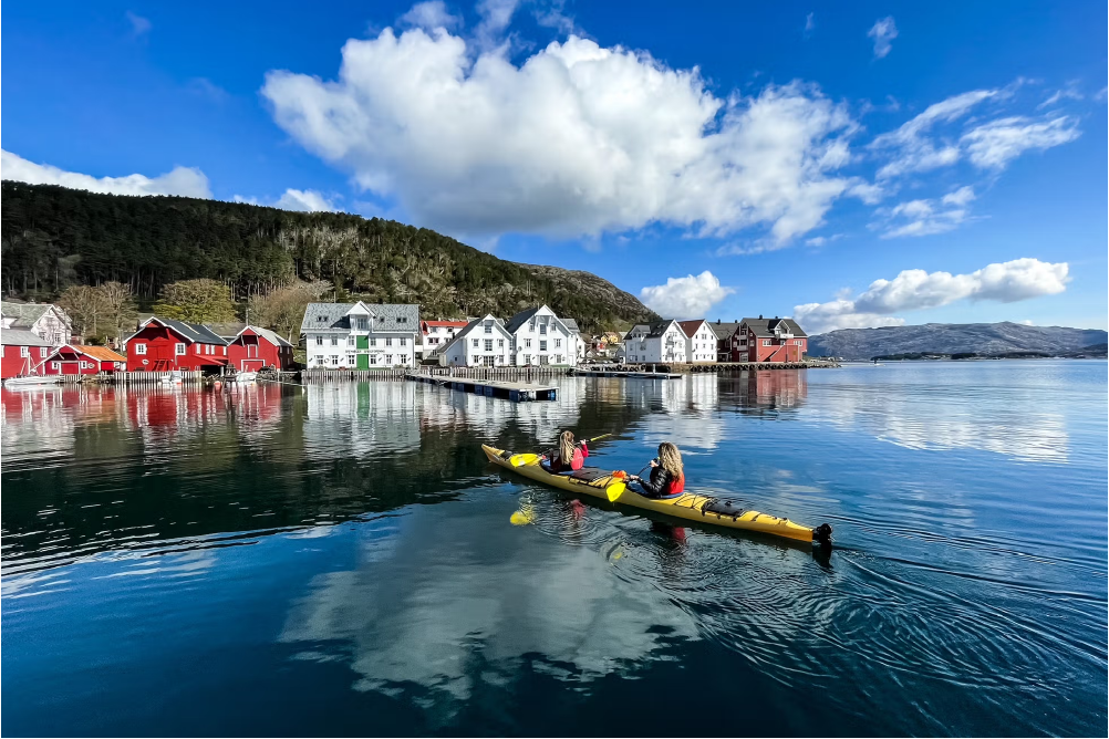 To personar i ein kayak padlar inn mot Kalvågs hus i typisk fiskevær-stil. Foto.