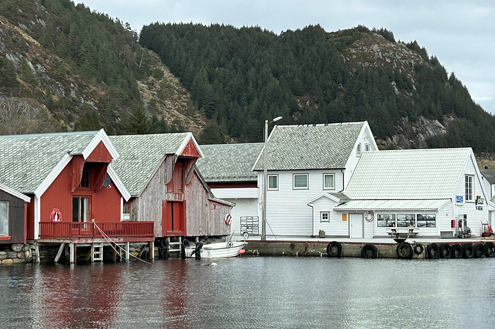 Raude naust og kvite hus sit på kaien. Det er ein kvit båt fortøydd til kaien. Kystlandskap i bakgrunnen. Foto.