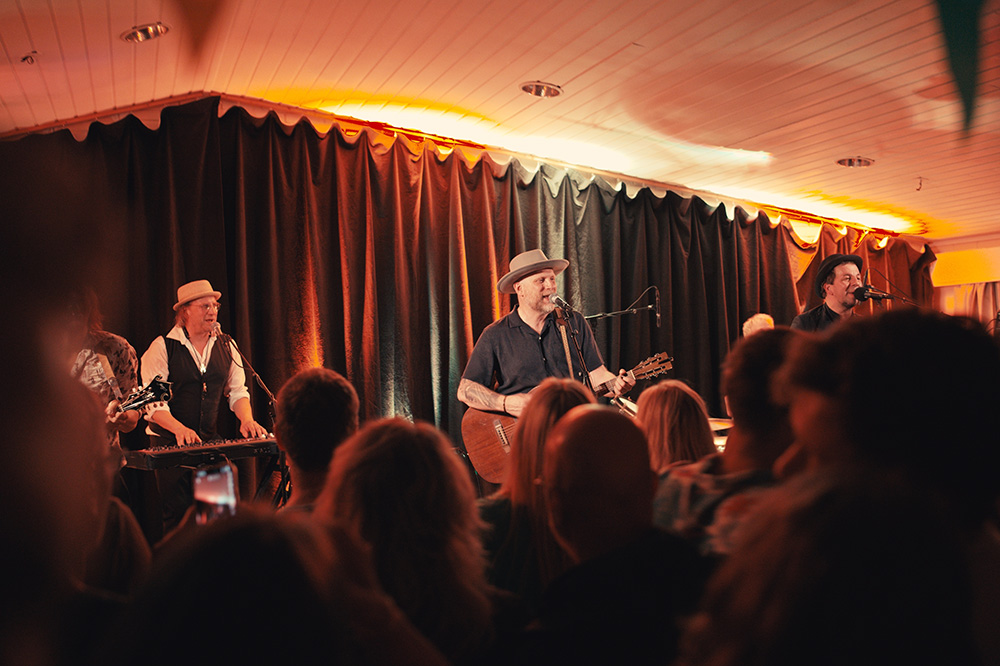 Mannleg band spelar på intim scene framfor eit publikum. Gitarane er akustiske og dei har på seg pork pie-hattar, altso flate hattar med tynn brem. Foto.