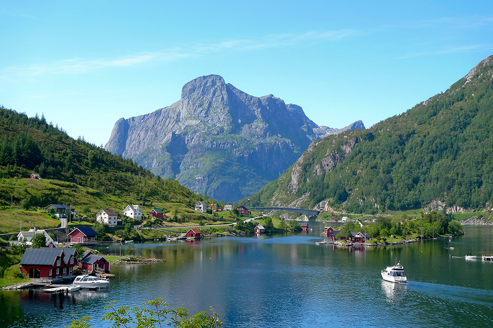 Den store fjellklippa Hornelen i bakgrunnen er ramma inn av eit landskap med fjord og fjell på kvar side. Det er ein liten båt som køyrer i retning av fjellet.